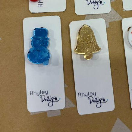 Set of Christmas-themed pins on cards with 'Rhyley Design' branding against a brown background with tree patterns.
