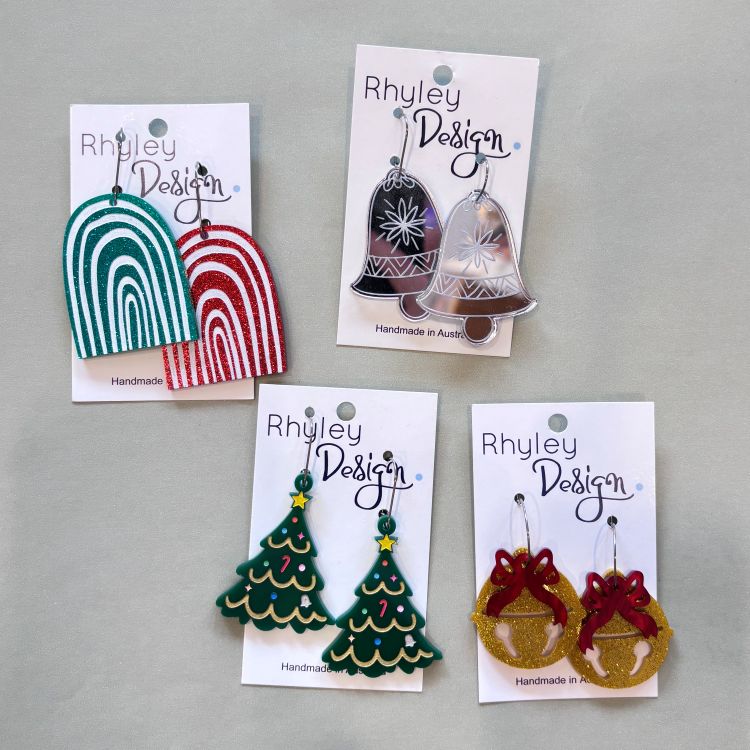 Four pairs of handmade earrings displayed on white cards against a light gray background. The designs include two arch-shaped earrings in teal and red, two bell-shaped earrings in silver and clear, two Christmas tree earrings in green, and two bell-and-crown earrings in gold and red.
