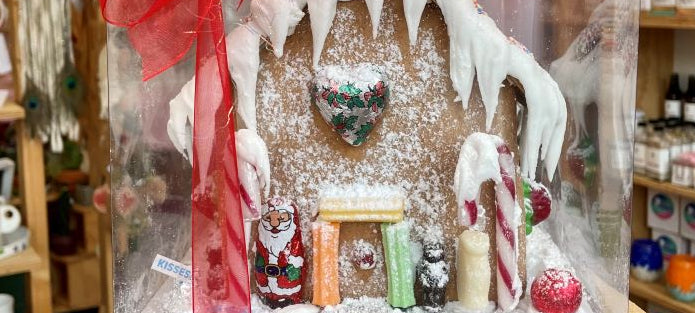 A decorated gingerbread house with a red bow on top, surrounded by various holiday-themed candies and decorations, set against a blurred store background.